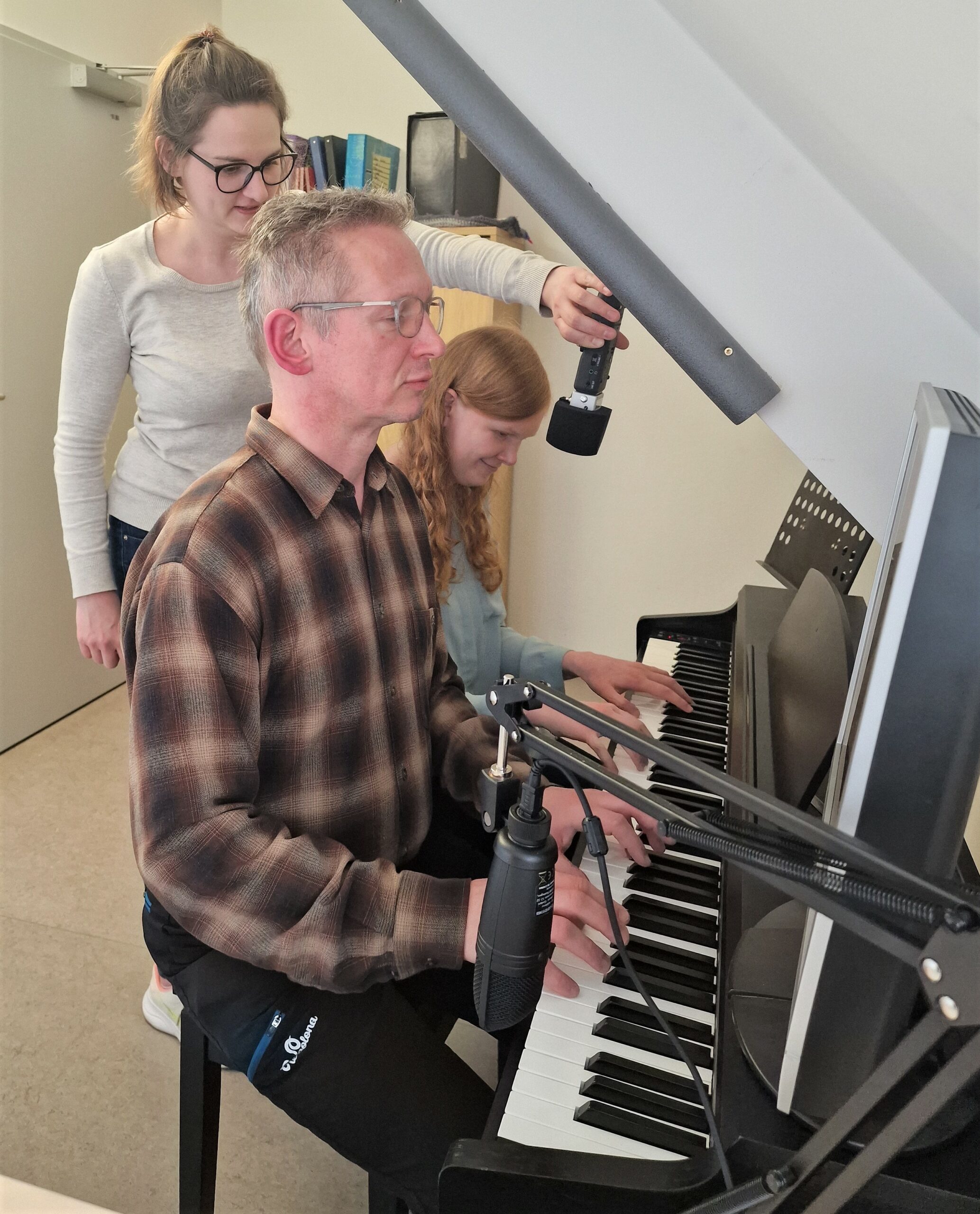 Im Raum steht auf der rechten Wandseite ein Klavier; am Klavier sitzen die Projektkoordinatorin und Probeschüler; dahinter steht die Interviewerin und hält ein Mikrofon zwischen den beiden